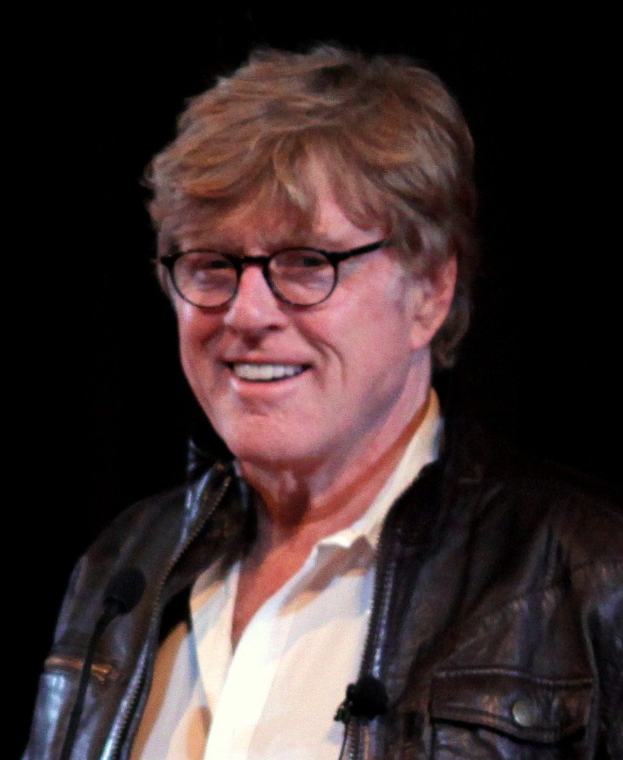 Robert Redford smiling in 2009, wearing glasses and a leather jacket at a public event